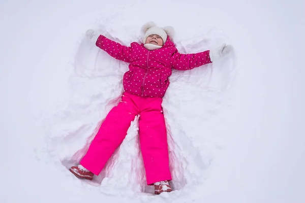 Hermosa niña hace un ángel de nieve en un parque, vista superior . — Foto de Stock