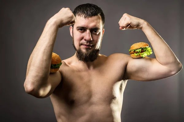 Mladý muž kulturista držení burger, nezdravé stravovací návyky. — Stock fotografie