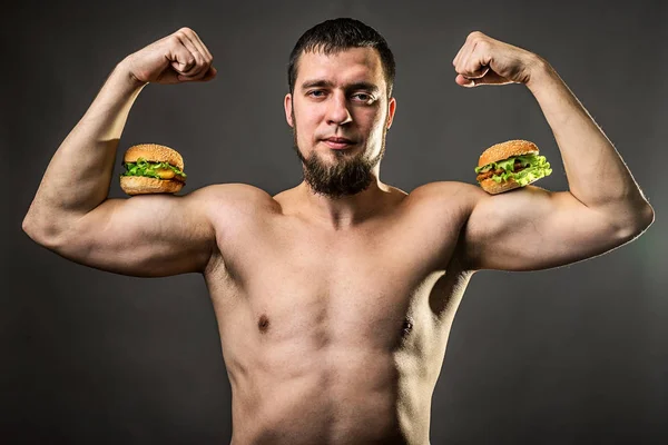 Hombre joven culturista sosteniendo hamburguesa, hábitos alimenticios poco saludables . — Foto de Stock