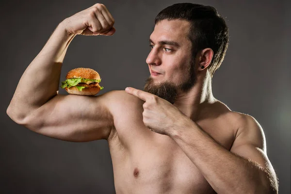 Hombre joven culturista sosteniendo hamburguesa, hábitos alimenticios poco saludables . — Foto de Stock