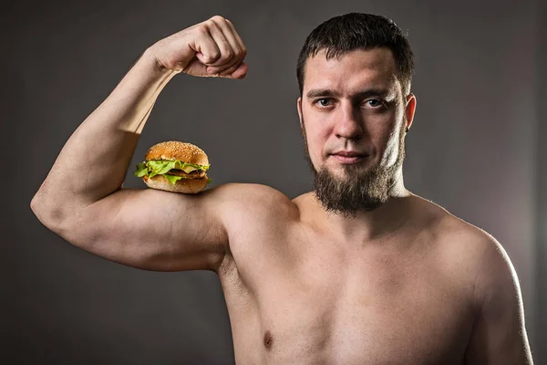 Hombre joven culturista sosteniendo hamburguesa, hábitos alimenticios poco saludables . — Foto de Stock