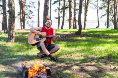 Genç adam kamp ateşi yanında otururken akustik gitar çalmak.