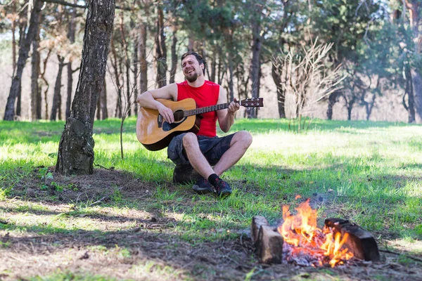 Jeune homme jouant de la guitare acoustique assis près de feu de camp . — Photo