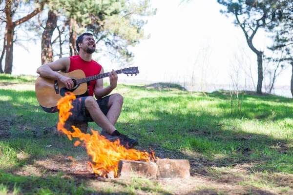 Jongeman akoestische gitaar spelen terwijl u zit in de buurt van kampvuur. — Stockfoto
