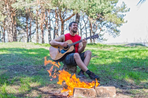 Genç adam kamp ateşi yanında otururken akustik gitar çalmak. — Stok fotoğraf