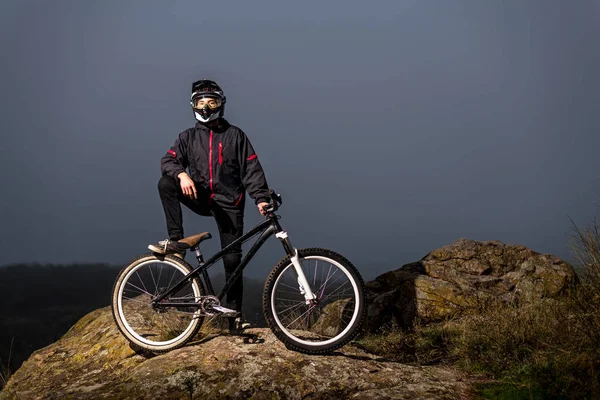 The rider in the full-face helmet and full protective equipment on the mtb bike stands on a rock. — Stock Photo, Image