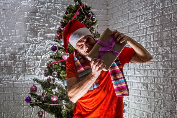 Um Papai Noel irritado e agressivo fica perto da árvore de Natal e mantém um presente de Ano Novo . — Fotografia de Stock