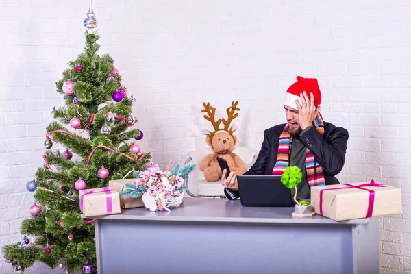 Uomo d'affari in giacca e cravatta e cappello da Babbo Natale che lavora su un portatile alla vigilia di Natale, uno stacanovista . — Foto Stock