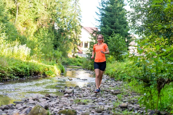 Chica en una camiseta naranja brillante corriendo a lo largo de un río de montaña . — Foto de Stock