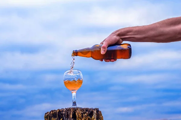 Una mano masculina vierte una bebida en un vaso sobre el fondo de un hermoso cielo . — Foto de Stock