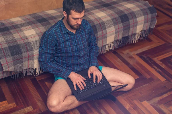 A bearded man is working on a laptop while sitting on the floor near the sofa. The concept of remote work, quarantine.