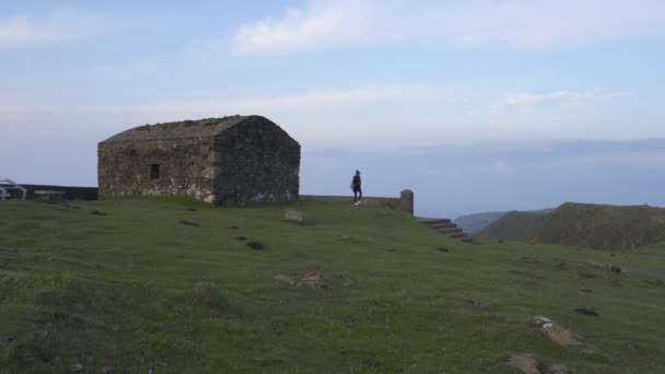 女性はスペインのVixia Herbeiraの緑の山の風景の日没で家の廃墟の近くを歩く — ストック動画