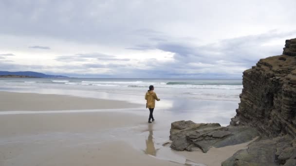 女性の女の子旅行者ラス カテドラレスの上を歩く素晴らしい風景ビーチ低潮で嵐の雨の日に黄色のジャケット — ストック動画
