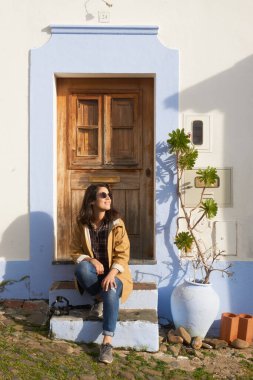 Sarı ceketli kadın turist, Portekiz, Mertola Alentejo 'da bitkisi olan güzel bir antika beyaz ve mavi evin merdivenlerine oturdu.