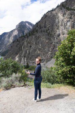 Elinde cep telefonu ve kamerasıyla kameraya bakan gülümseyen genç kadın Wells Gray eyalet parkı, British Columbia, Kanada