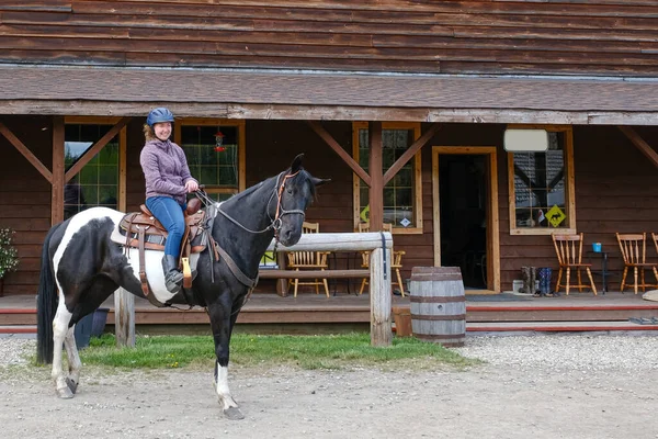 A young woman riding a black and white horse in front of a wooden house, Banff national park — Stock Photo, Image