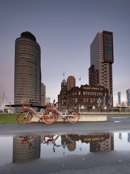 Rotterdam - 2019. február 12.: Rotterdam, Hollandia. Hotel New York-ban. Két piros kerékpár parkolt a szálloda előtt, a szálloda és a kerékpárok tükröződik a víz az úton, éjjel a — Stock Fotó