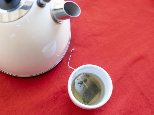 Electric kettle with boiling water for the herbal tea. Tea bag in a white tea cup on a red table, Selective focused on tea cup
