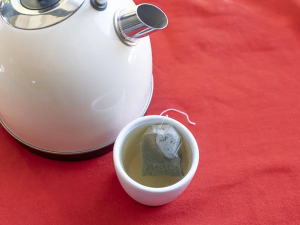 Electric kettle with boiling water for the herbal tea. Tea bag in a white tea cup on a red table, Selective focused on tea cup