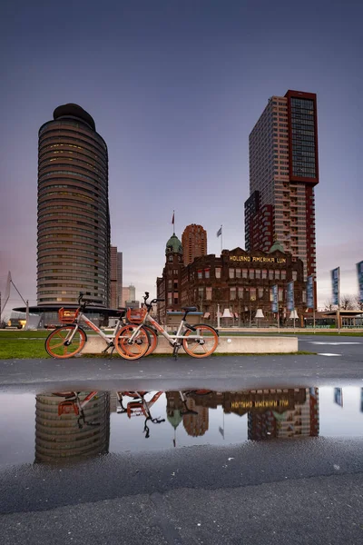 Rotterdam - 12 February 2019: Rotterdam, The Netherlands. Hotel New York. Two red bicycles parked in front of the hotel, the hotel and the bicycles reflected in the water on the footpath, at Night on — Stock Photo, Image