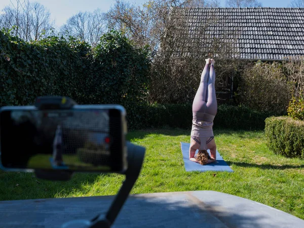 Bahçede yoga egzersizleri yapan genç bir kadın, yoga pozları tripoda cep telefonuyla çekiliyor.