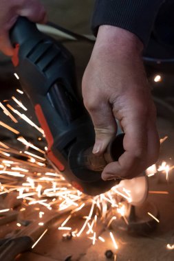 Endüstriyel atölyede kıvılcımlar uçuşurken elektrikli değirmen kullanan, bahçe tezgahının metal kısmında çalışan bir adamın ellerine ön bakış..