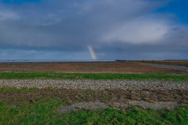 Bulutlu koyu mavi gökkuşağı. Çimenler ve taşlar ön planda. Uzaktaki insanlar. Hollanda: Waddensea. Unesco Dünya Mirası.