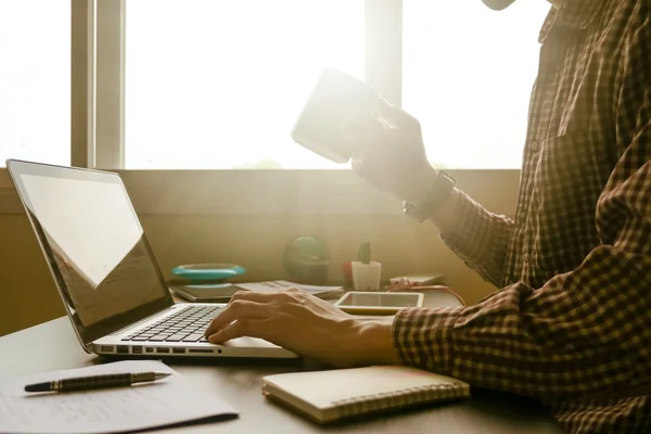 Un hombre sosteniendo una taza de café y trabajando en el portátil en su escritorio de oficina, tonificado con la luz del sol . — Foto de Stock