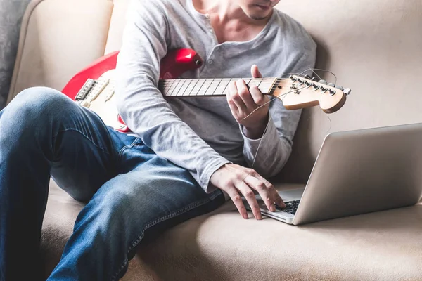 Casual genç adam evde kanepede dizüstü bilgisayar ile gitar çalmak.