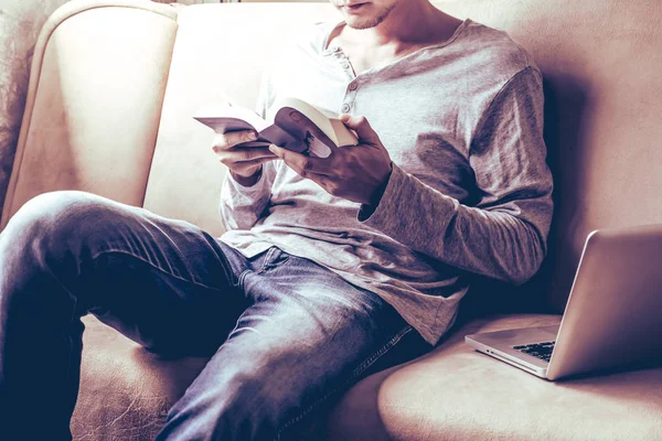 Casual young man reading book on sofa at home.