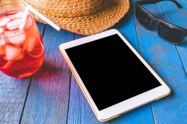 Şapka, güneş gözlüğü ve kırmızı mavi boyalı ahşap tahta arka plan üzerinde kokteyl ile tablet.