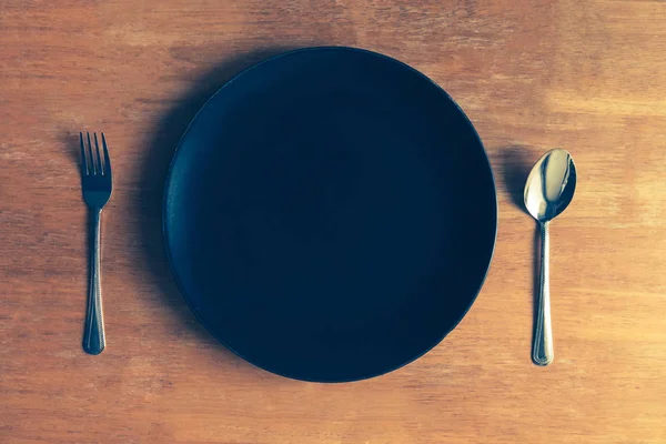 Draufsicht auf leeren schwarzen Teller mit Löffel und Gabel auf Holztisch — Stockfoto