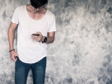 Genç Beyaz t-shirt ve kot giyen, akıllı telefon tutarak ve gri grunge arka plan üzerinde duran adam. Metin için boş alan