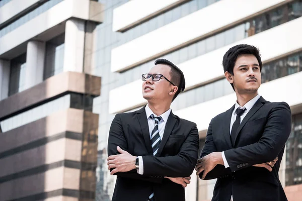Porträtt av två unga asiatiska affärsmän i svart kostym stående med kontorsbyggnad bakgrund — Stockfoto