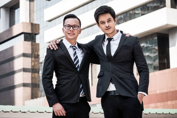 Retrato de dois jovens empresários asiáticos de terno preto em pé com fundo de prédio de escritórios — Fotografia de Stock