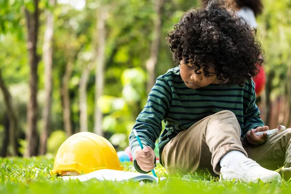 Un petit garçon écrit sur un cahier assis sur de l'herbe verte dans un parc — Photo