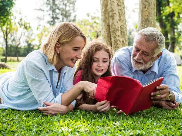 Atividades na família, pai, mãe e filha lendo um livro enquanto estabelece na grama verde em um parque — Fotografia de Stock