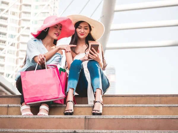 Dos hermosas mujeres jóvenes sentadas en las escaleras y utilizando el teléfono inteligente después de disfrutar en las compras —  Fotos de Stock