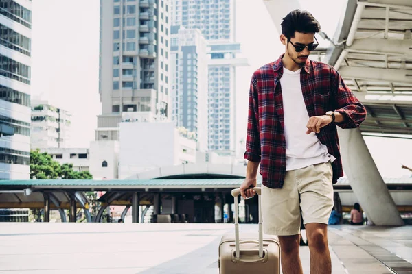 Young Asian tourist with luggage looking at his watch while walking on city street. Travel concept