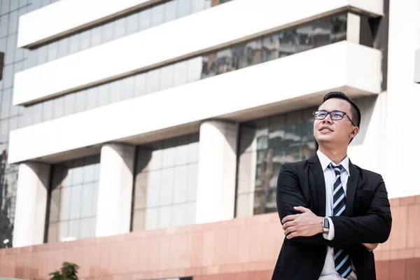 Portrait of young Asian businessman in black suit standing with office building background. Free space for text — Stock Photo, Image
