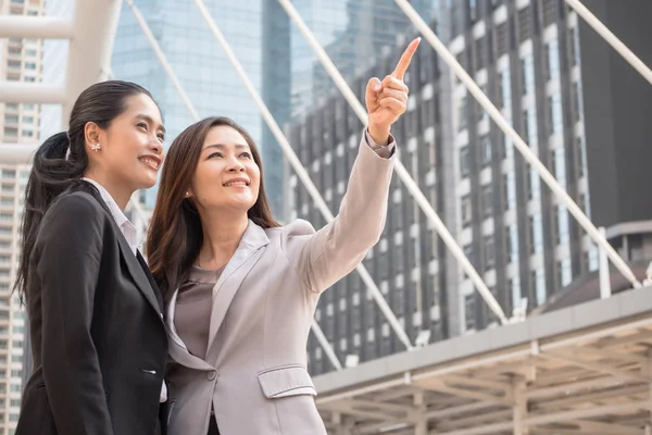 Two Asian business women pointing and smiling with urban scene background. Purpose and success concept