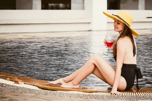 Młoda kobieta w strój kąpielowy z kapelusz i napoju siedząc na brzegu basenu. — Zdjęcie stockowe