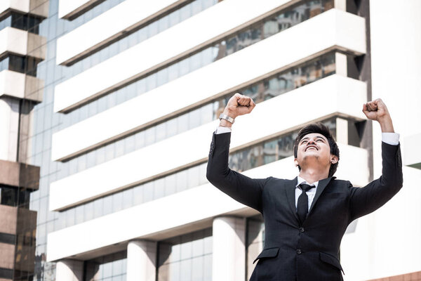 Portrait of young Asian businessman in black suit standing with office building background. Free space for text