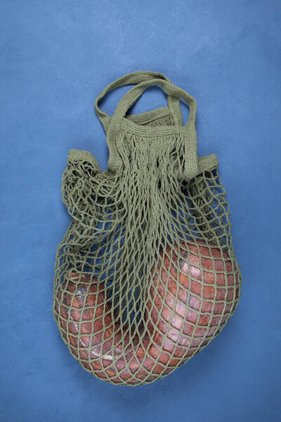 Funny, curved pumpkin butternut of the Guitar variety in a string bag on a blue background. Concept - reduction of food waste. Eating ugly vegetables and fruits. Vertical orientation. Copy space.