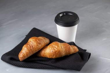 İki taze kruvasan koyu bir peçetenin üzerinde yatıyor. Yakınlarda beyaz bir kağıt bardak ve kahve var. Seçici odaklanma. Boşluğu kopyala.