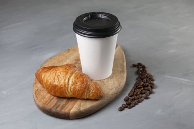 Taze kruvasan ve beyaz kağıt bardakta kahve gri arka planda tahta bir kesme tahtasının üzerinde. Yakınlarda birkaç kahve çekirdeği var. Seçici odak.