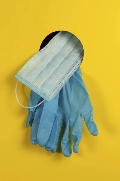 Medical concept. Disposable gloves and face mask through a hole on a yellow background as a means of protection against coronavirus infection nCov-2019. Closeup.