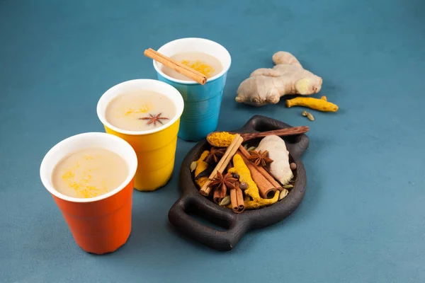 Çok renkli seramik bardaklarda Masala çayı / Masala çayı. Altın süt ve diğer Hint içecekleri için baharatlı ahşap tabak. Mavi beton arka plan. Boşluğu kopyala.