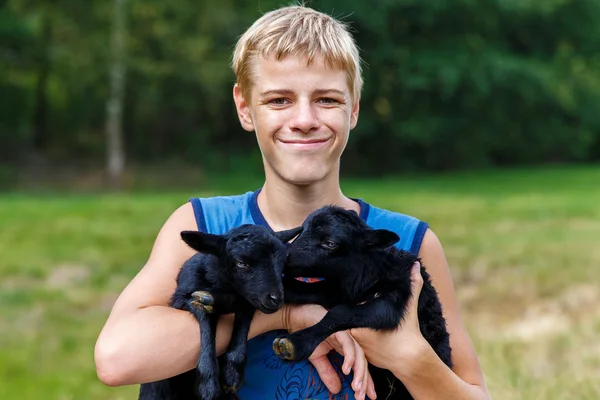 Happy teenager boy holding little black baby goats in the meadow — Stock Photo, Image