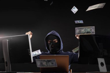 pleased young hacker man in mask sit in space full of money